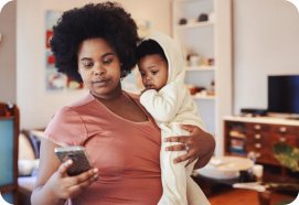 A mother holding her baby while checking her phone.