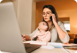 A mother working at a laptop with her baby beside her.